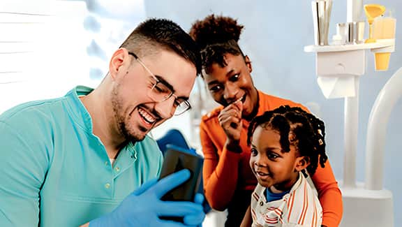 A dental professional connects with his young patient, while the child’s mother looks on with joy.