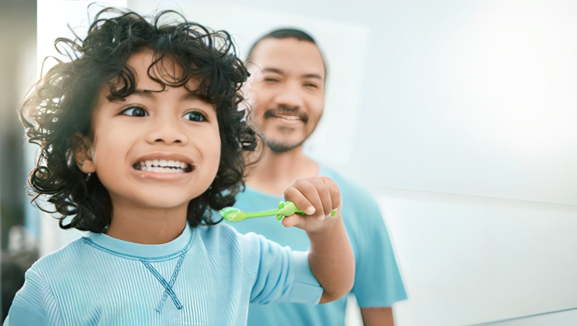 A young boy gazes at his own smile while brushing his teeth, and his father looks on with pride and amusement in the background.