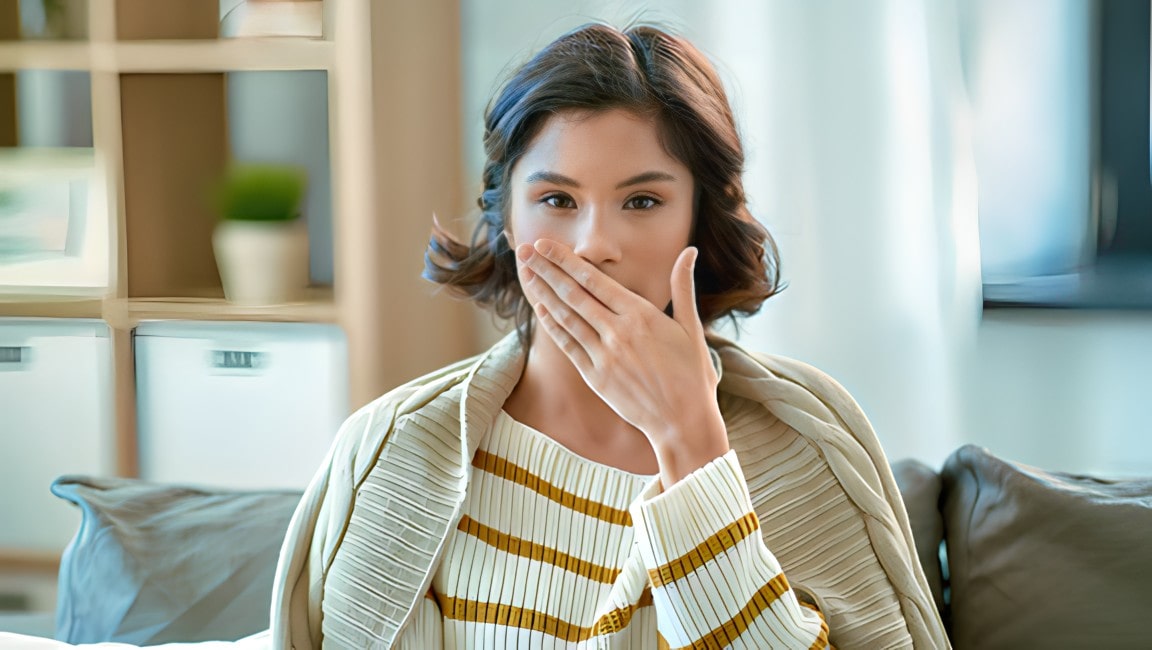A young woman, covering her mouth with one hand, as she realizes she may have bad breath.