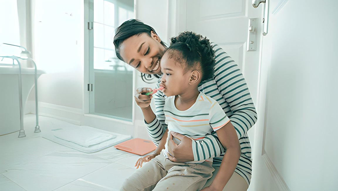 A young mother, hold her daughter in her lap in a bathroom, and she assists in brushing her teeth.