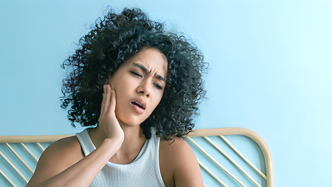 A young latina female with curly hair, touching her cheek in pain from a toothache, while sitting on a bed at home.