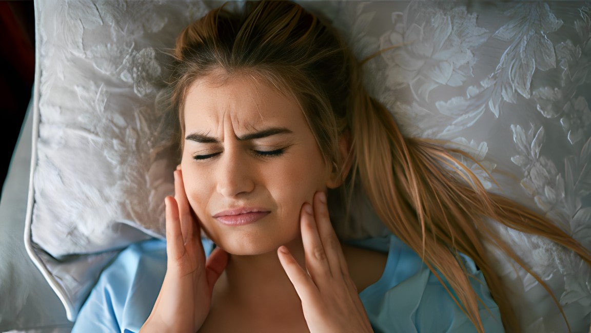 A young woman laying in bed, experiencing pain from grinding their teeth